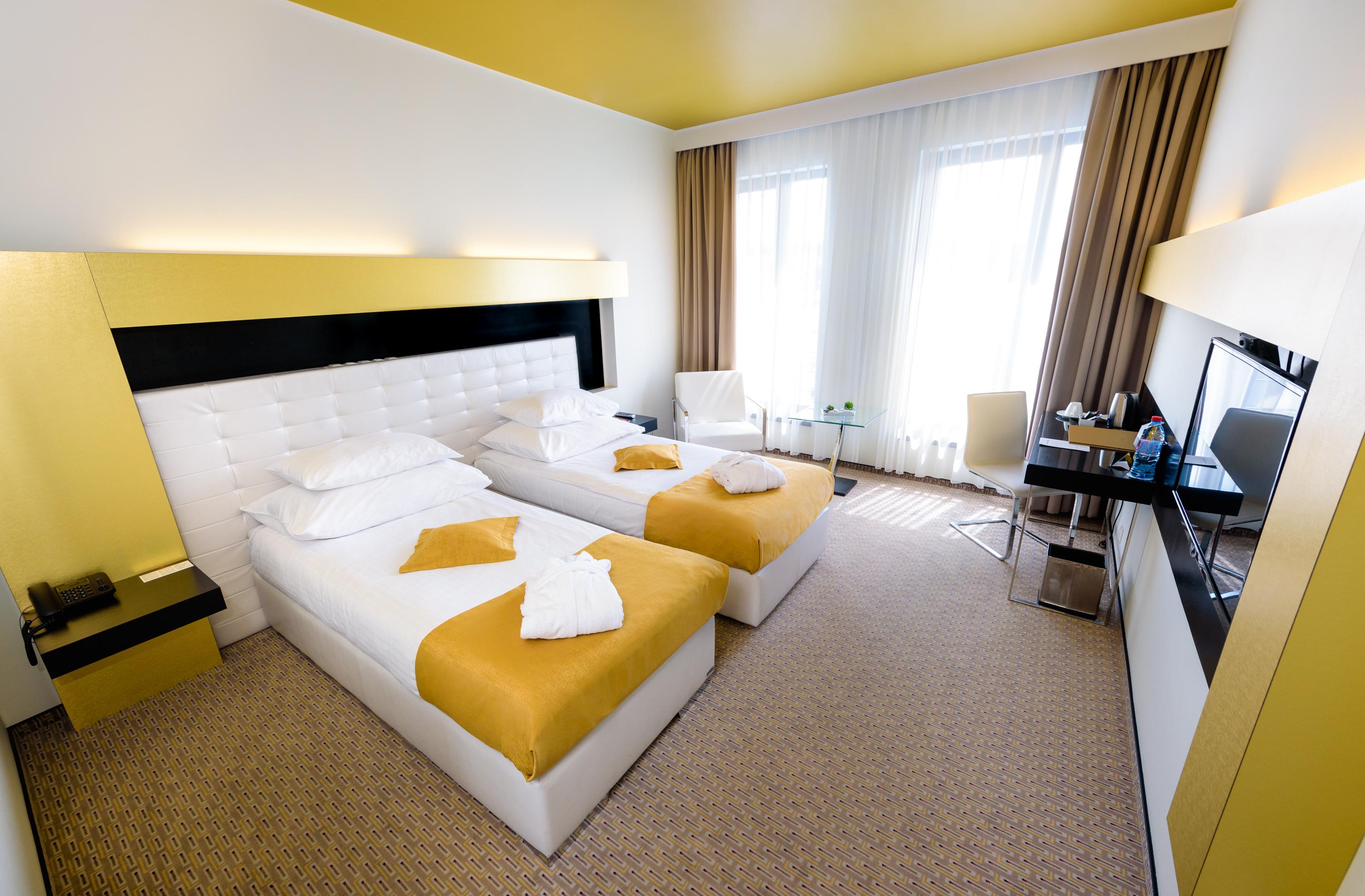 Grandior Hotel Praha Eksteriør bilde The photo shows a modern hotel room featuring two twin beds with white bedding and yellow decorative blankets. The room has a bright, minimalist design, with a yellow ceiling and light-colored walls. Large windows allow natural light to fill the spac